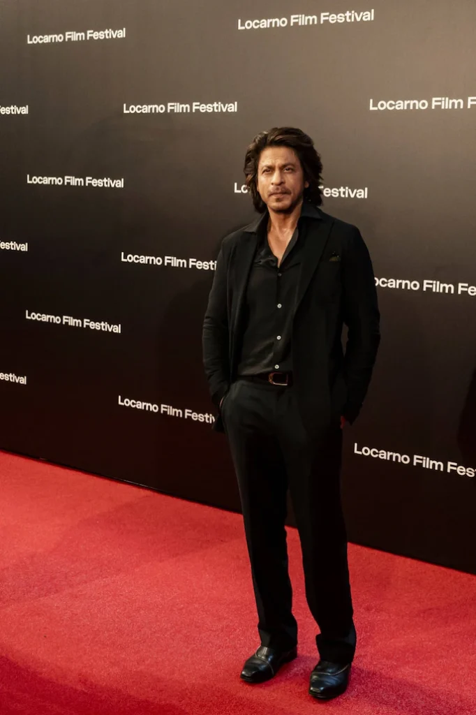 Shah Rukh Khan stands against a backdrop featuring the text ‘Locarno Film Festival’ repeated in a pattern. He is dressed in a black suit with a buttoned shirt and has his head bowed slightly,