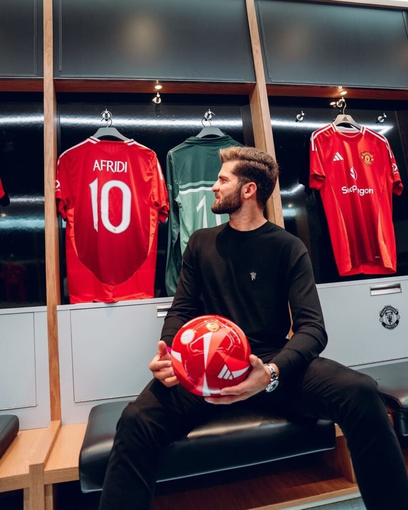 Shaheen Shah Afridi is sitting in a locker room, holding a red soccer ball with a Manchester United logo. The person is wearing a black long-sleeve top with a small white logo on the chest and dark pants. Behind the individual, there are three soccer jerseys hanging: two red ones with ‘AFRIDI’ and the number ‘10’ printed on them, and one green goalkeeper jersey in between. The locker room has wooden benches and lockers, with a white towel draped over one of the lockers.