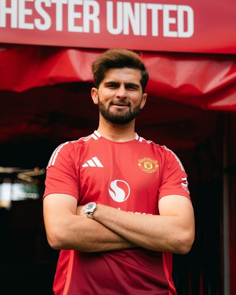 Shaheen Shah Afridi stands confidently with his arms crossed in front of a ‘Manchester United’ banner. He is wearing the team’s red home jersey with a white logo on the right chest and the club’s emblem on the left. His face is obscured for privacy, and he is wearing a watch on his left wrist.