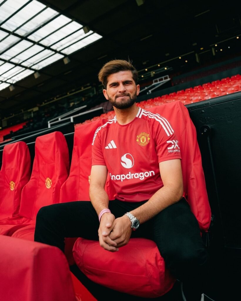 Shaheen Shah Afridi sits on a red stadium seat, wearing a red sports jersey with the word ‘Snapdragon’ and a logo on it, along with dark trousers. His face is obscured by a brown pixelated rectangle for privacy. The setting appears to be a large sports stadium with several empty seats around him.