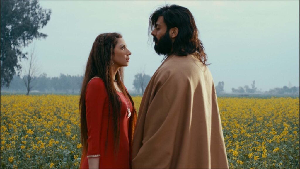 Image features Mahira Khan and Fawad Khan standing in a field of yellow flowers. Both are facing away from the camera, looking into the distance. Mahira Khan is wearing a red outfit, while Fawad Khan is clad in a beige garment.