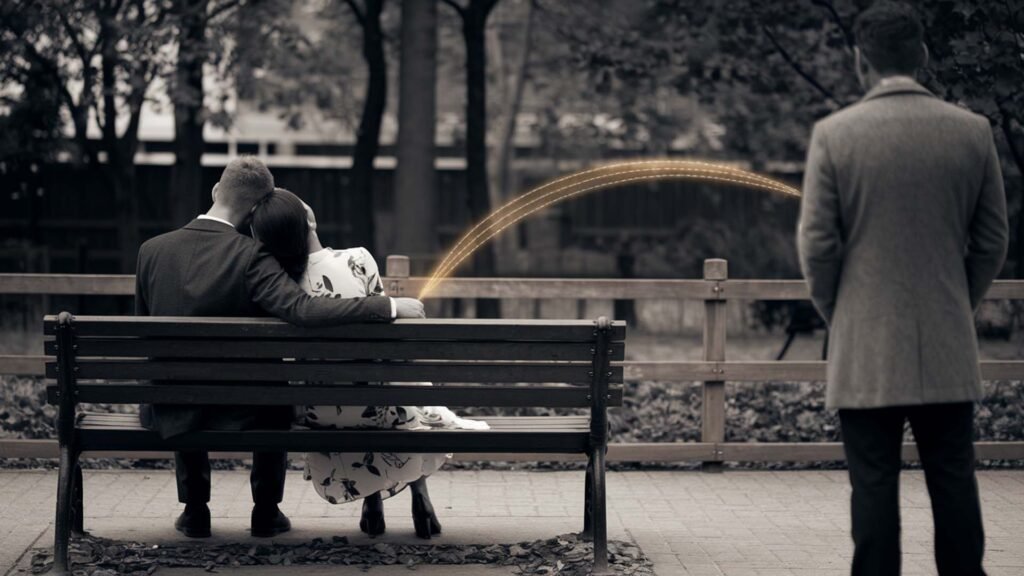 The image depicts three individuals in an outdoor setting with trees and a walking path. In the foreground, two individuals are seated closely on a bench; one has their head resting on the other’s shoulder, suggesting a moment of closeness or comfort. The third individual stands at a distance with their back to the camera, appearing to observe the seated pair.