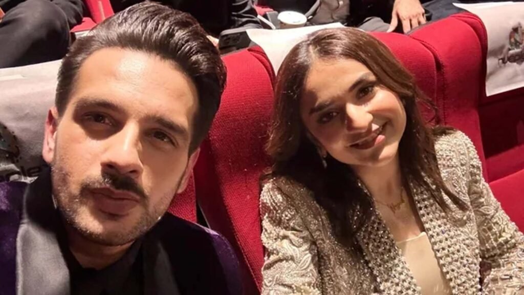 The image shows Yumna Zaidi and Usman Khan, both Pakistani actors, seated next to each other at an event. Yumna Zaidi is wearing a gold and beige patterned outfit, while Usman Khan is dressed in a dark-colored suit. They are in an indoor setting that resembles a theater or auditorium, indicated by the red seats in the background.
