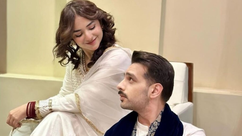 The image shows Yumna Zaidi and Usman Khan, both Pakistani actors, standing together at a formal event. Yumna Zaidi is wearing a traditional outfit with intricate embroidery, while Usman Khan is dressed in a suit. They are both smiling and looking at the camera, with a backdrop featuring event branding behind them.