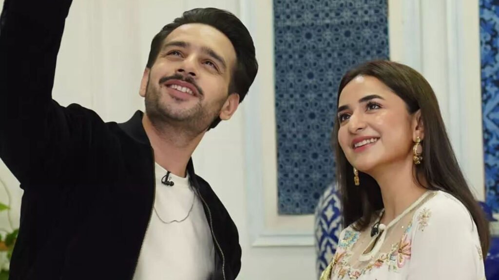 The image shows Yumna Zaidi and Usman Khan, both Pakistani actors, standing side by side. Yumna Zaidi is wearing a white outfit with floral patterns and earrings, while Usman Khan is dressed in a black outfit. They are positioned against a background featuring a blue patterned design.