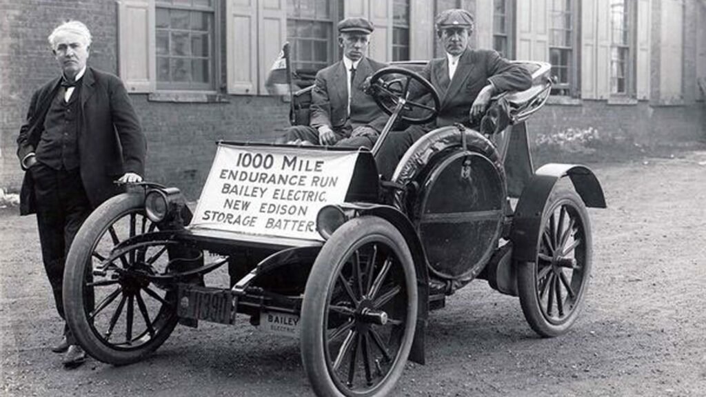 Three individuals seated in an early 20th-century electric car, participating in a 1000-mile endurance run to showcase the capabilities of the Edison storage battery.
