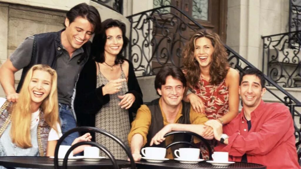 Group of six friends gathered around a table outside Central Perk, celebrating the 30th anniversary of Friends, with coffee cups and laughter evident despite