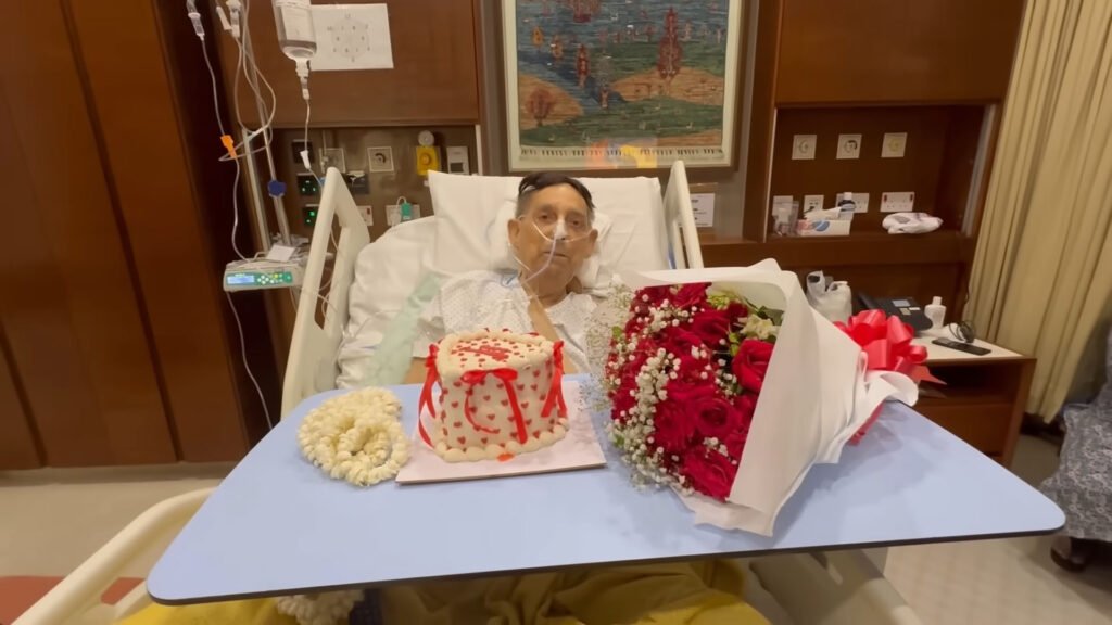 Shagufta Ejaz’s husband, Yahya Siddiqui, seated in a hospital bed with celebratory items including a cake and bouquets.