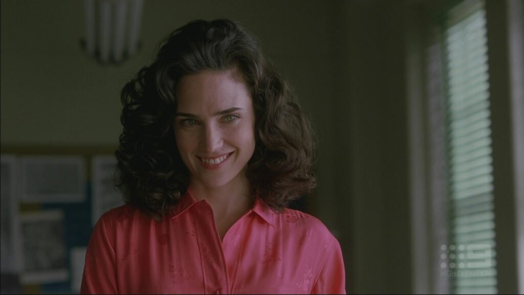 Image of a young Jennifer Connelly look-alike, long dark hair, wearing a pink shirt, standing in a room with green blinds.