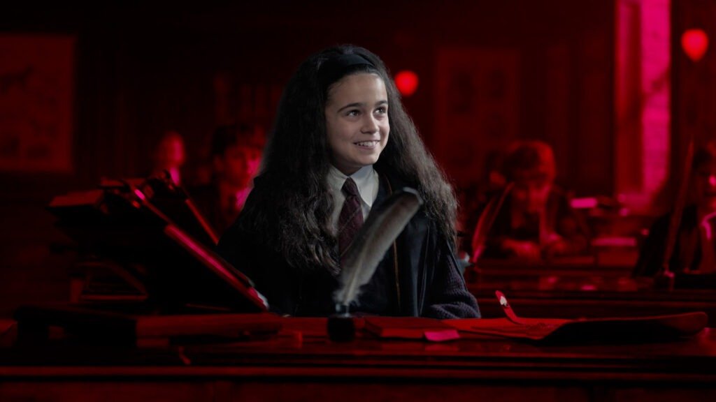 No Cap, the Harry Potter Trailer Just Reset the Entire Wizarding World 3 Close-up of young actress Arabella Stanton playing Hermione Granger. She is smiling while seated at a wooden desk in a dimly lit, red-hued classroom, wearing a traditional Hogwarts school uniform and tie with a large feather quill in the foreground.