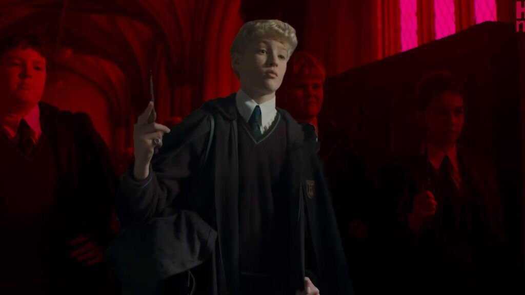 No Cap, the Harry Potter Trailer Just Reset the Entire Wizarding World 6 Actor Lox Pratt as Draco Malfoy, depicted as a young boy with platinum blonde hair, wearing a Slytherin school uniform and cloak. He is in a dimly lit Hogwarts stone corridor, illuminated by a strong red ambient light from the right. He holds up a wand with a distinctive silver ring on his finger and carries a travel bag. Two larger, slightly out-of-focus background actors representing Crabbe and Goyle flank him.