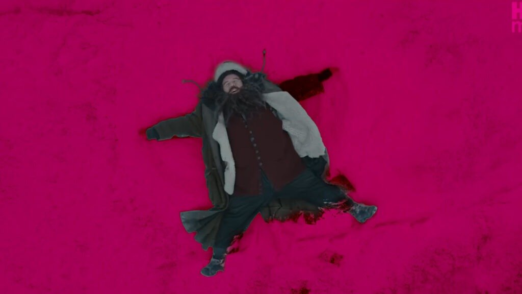 No Cap, the Harry Potter Trailer Just Reset the Entire Wizarding World 4 Actor Nick Frost, playing Rubeus Hagrid, lies on his back in a thick layer of pink-tinted snow, arms and legs spread wide as if making a snow angel. He is wearing Hagrid's iconic heavy brown overcoat, a fur-lined hat, and a messy, dark beard, with a joyful expression on his face.