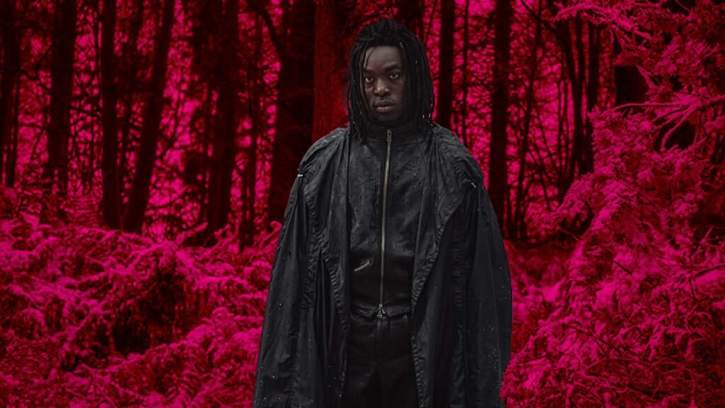 No Cap, the Harry Potter Trailer Just Reset the Entire Wizarding World 5 Actor Paapa Essiedu portraying a reimagined Severus Snape. He is wearing a long, layered black cloak and leather-style jacket with his hair in dark locs. He stands in a stark, snow-covered forest under a deep magenta and pink lighting grade, looking directly at the camera with a stern, intense expression.
